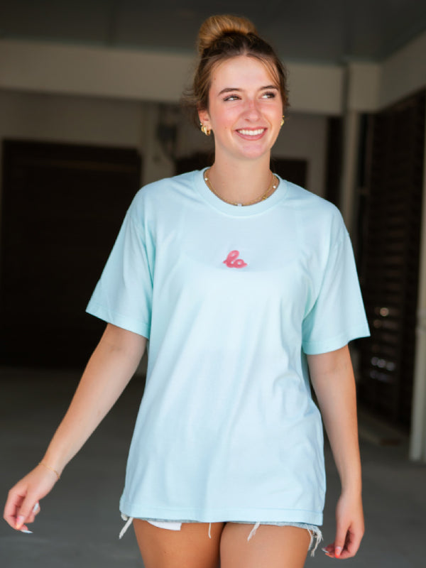 Close-up of embroidered logo on mint green tee, layered necklaces